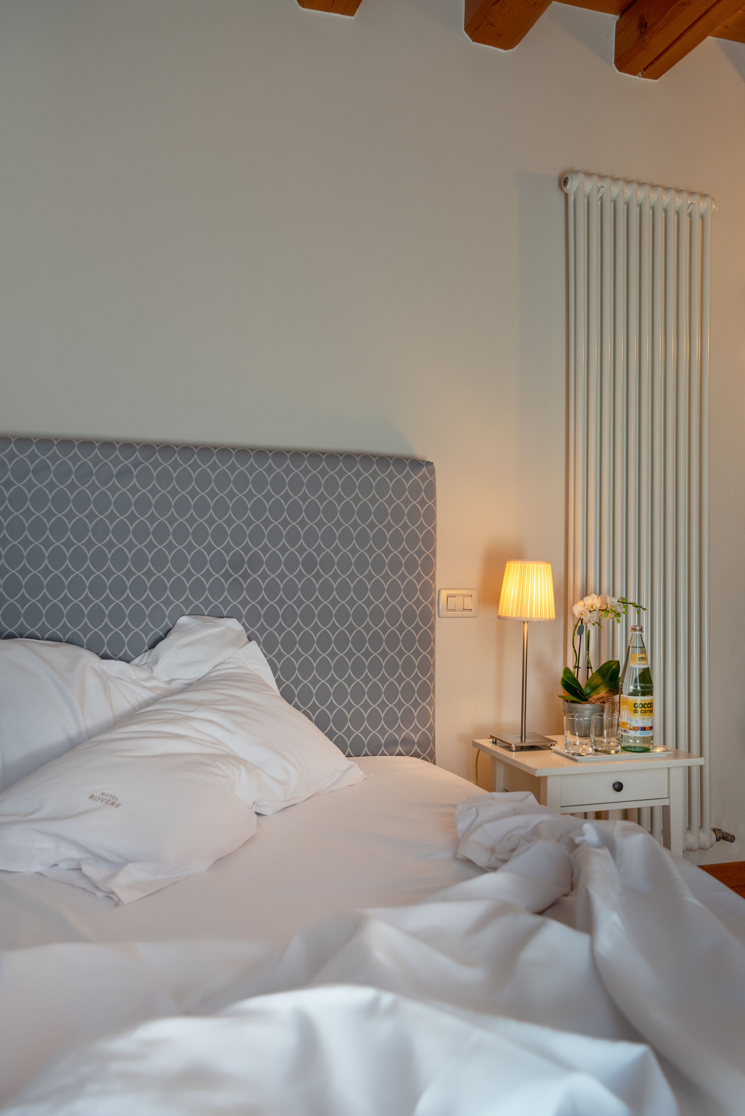 Una cama bien hecha con ropa blanca, una mesita de noche con lámpara y un radiador.