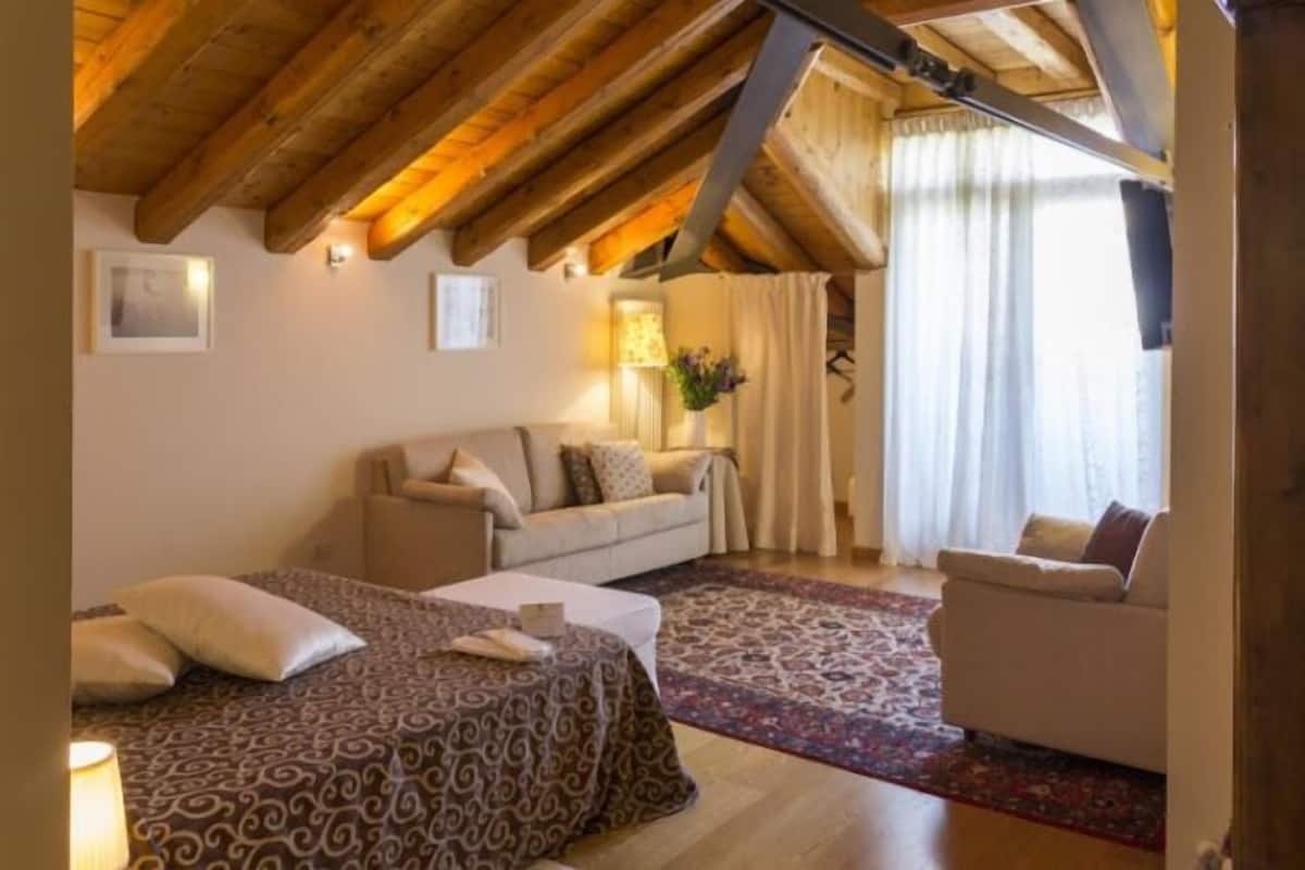 A cozy room with a bed, a sofa, and a small table. There is a window with curtains and a lamp on the wall.