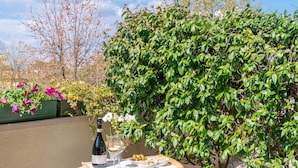 Un patio con una mesa preparada para dos personas, una botella de vino y una copa. Se ven sillas y una pequeña planta al fondo.