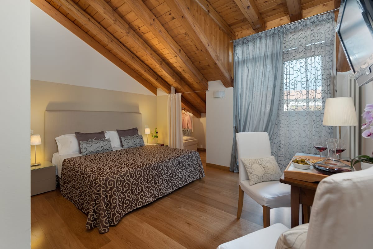 A bedroom with a wooden ceiling, a bed with patterned bedding, a bedside table with lamps, and a window with patterned curtains.