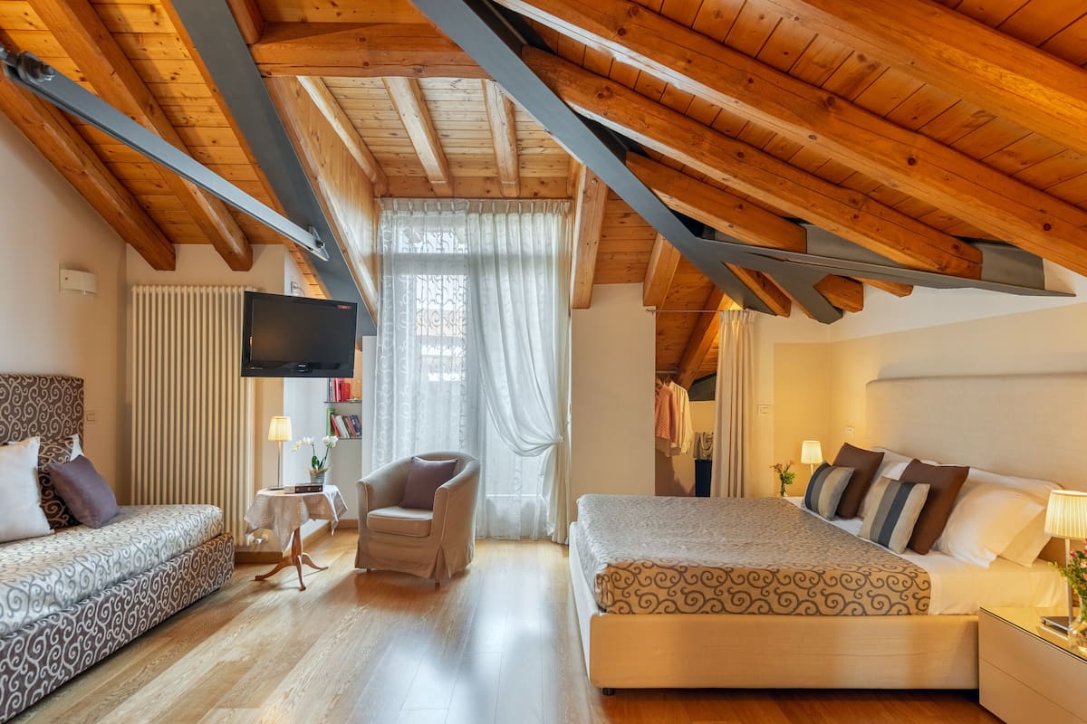 A bedroom with a wooden ceiling, a bed, a sofa, a chair, a TV, and a window with curtains.