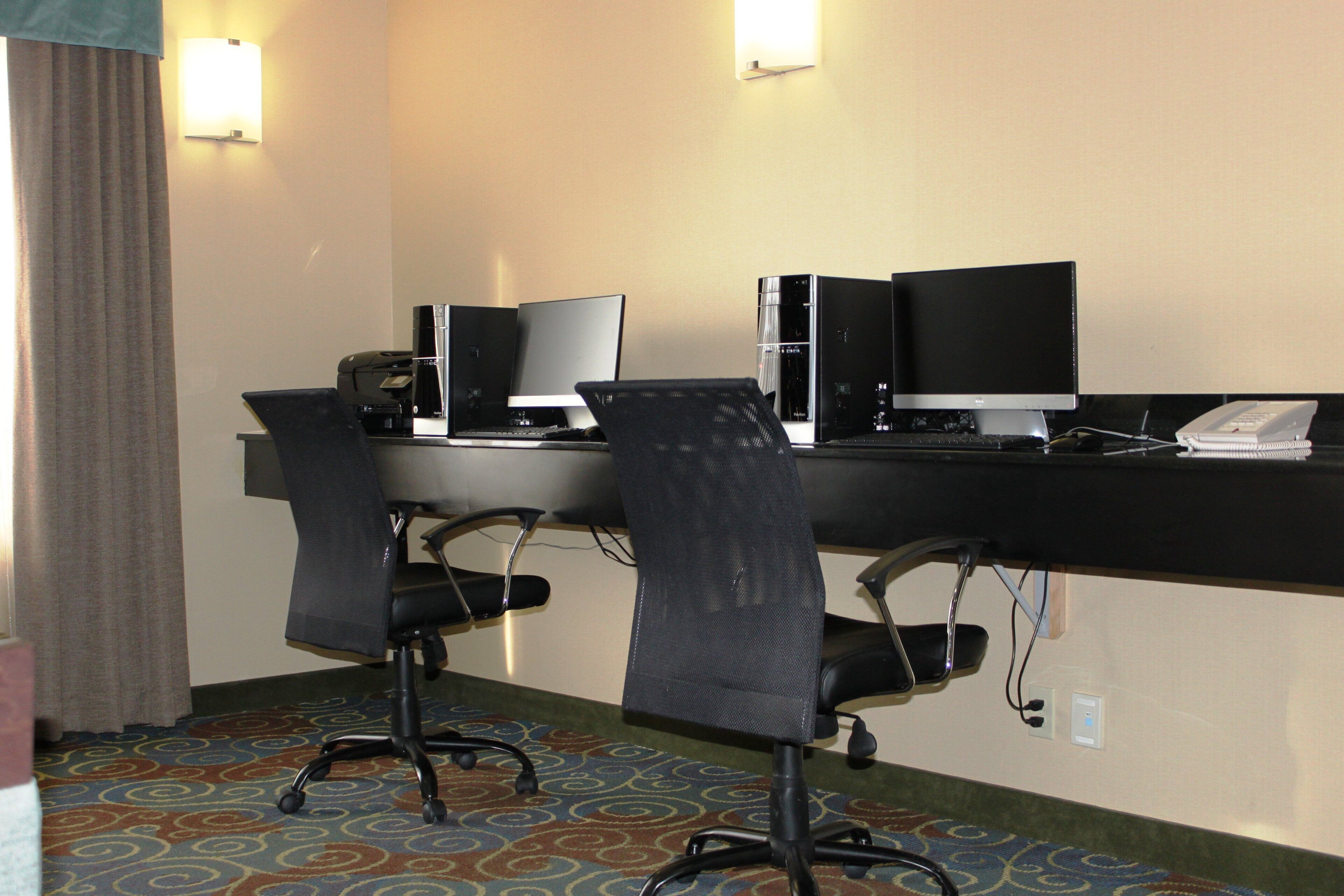 A hotel room with a desk, two chairs, a computer, a phone, and a lamp.