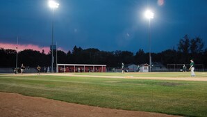 Een honkbalveld in de schemering met lichtmasten, een dug-out en spelers.