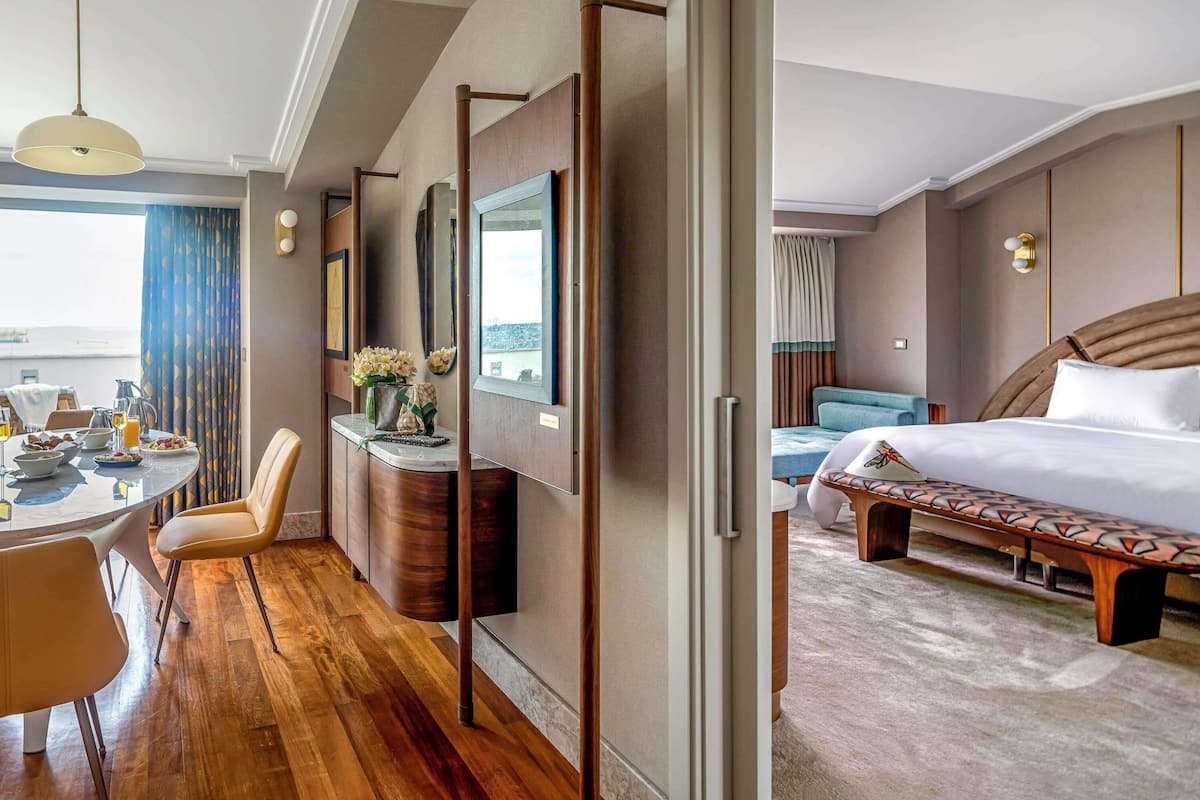 Habitación de hotel con cama, escritorio con silla, mesa de comedor con sillas, vista a la ciudad y un ventanal grande.