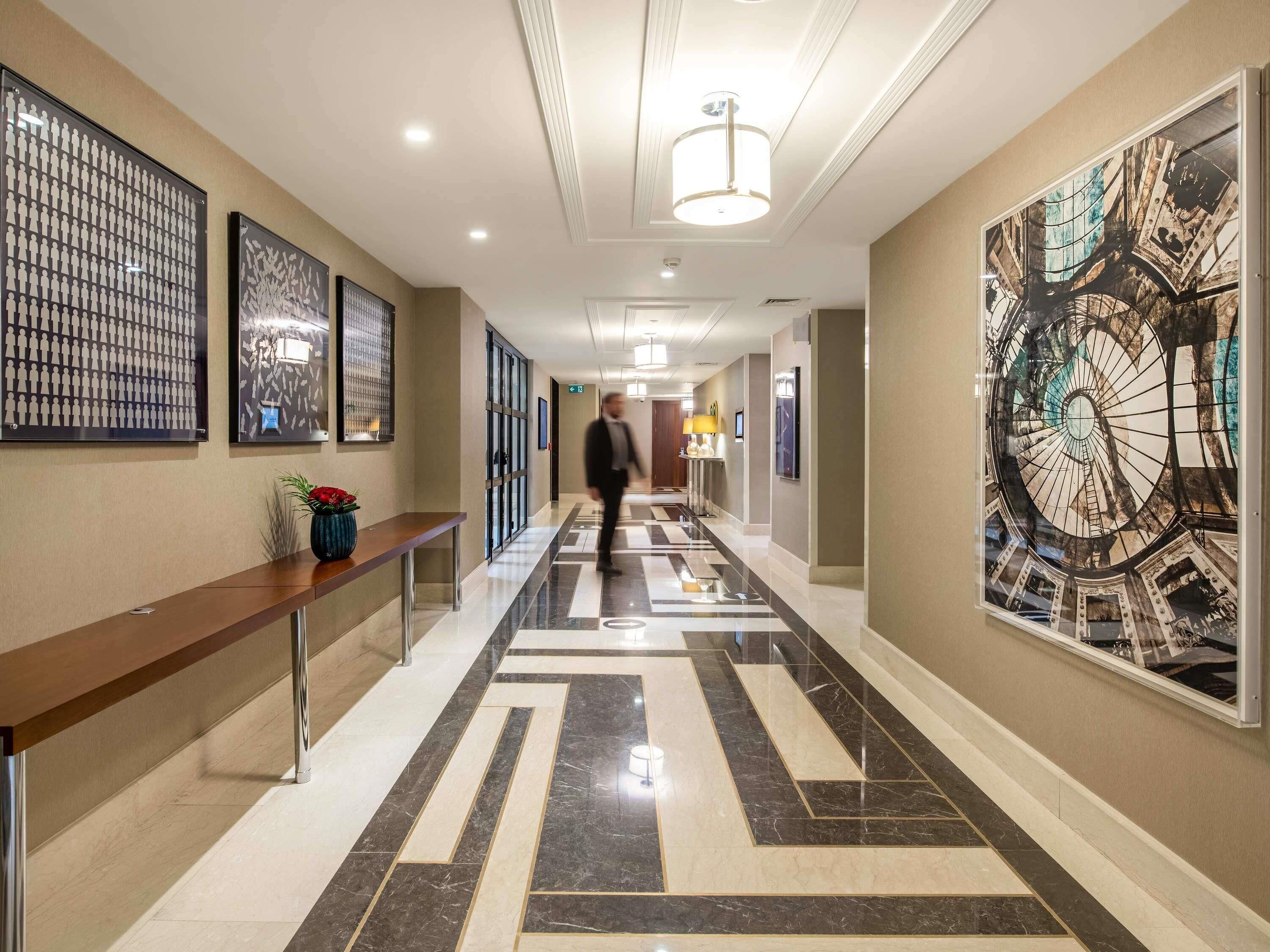 A hotel corridor with a marble floor, framed artwork on the walls, and a bench with a vase of flowers.