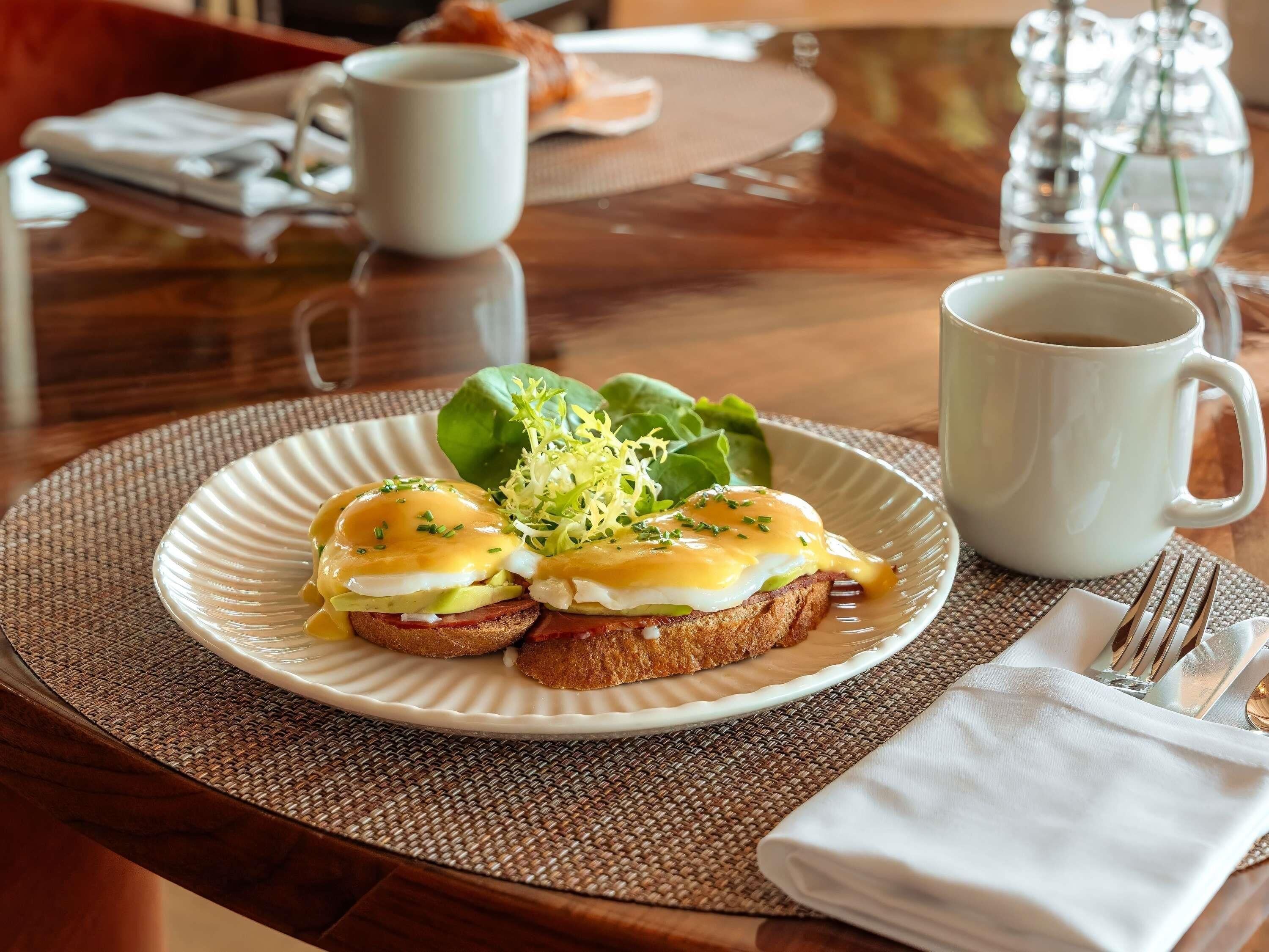 Un desayuno con huevos Benedict en un plato, una taza de café y una servilleta con cubiertos.