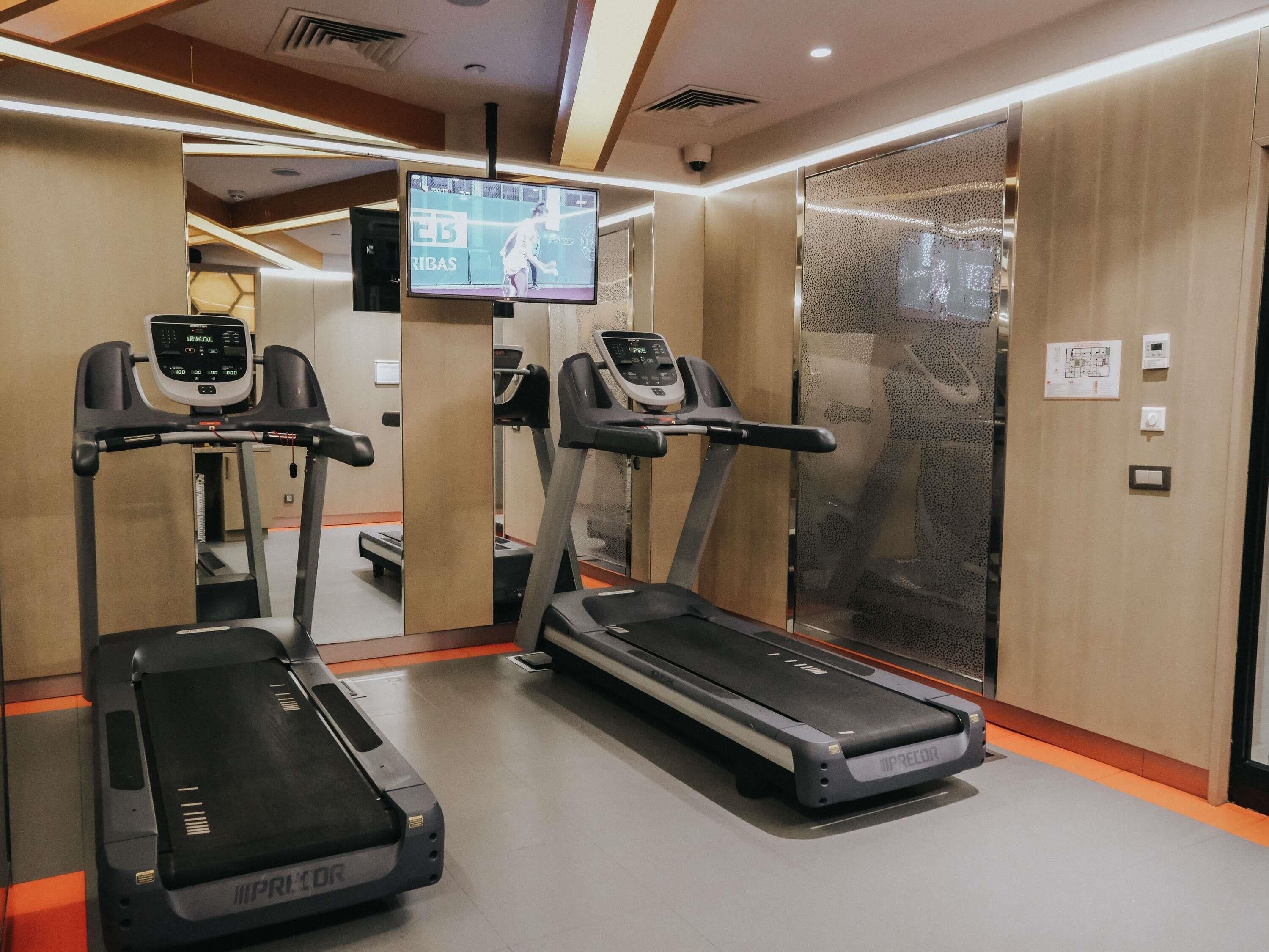 A gym with treadmills, a mirrored wall, and a television screen displaying a tennis match.