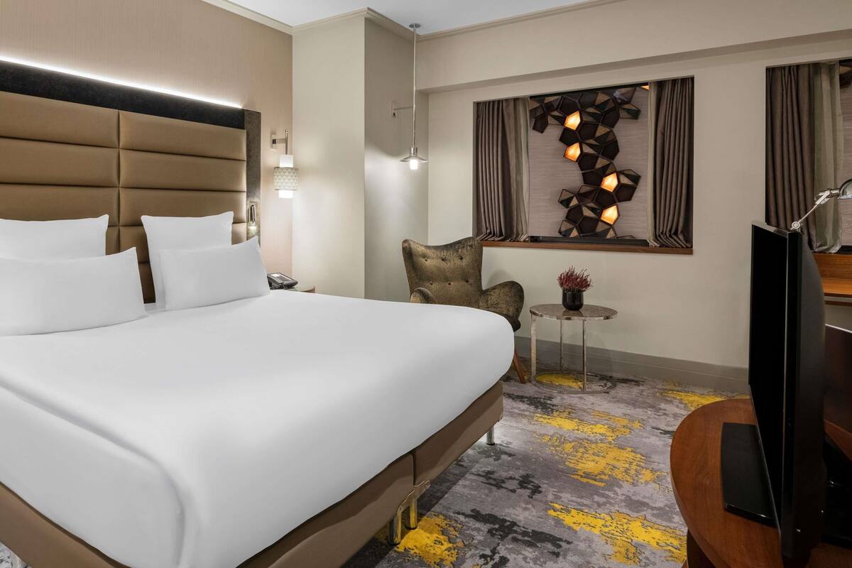 Een hotelkamer met een groot bed, een tafeltje, een stoel, een televisie en een decoratief kunstwerk aan de muur.