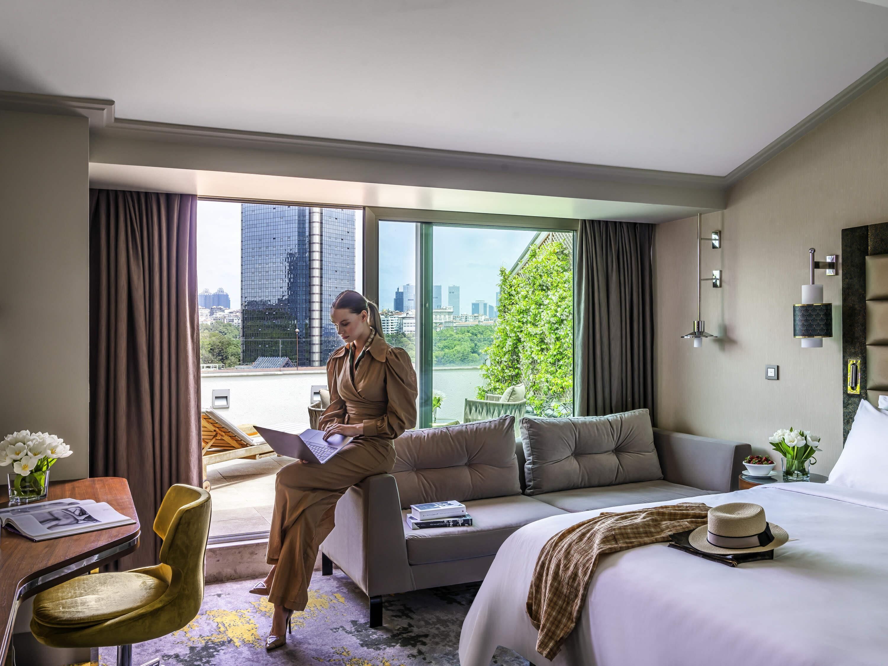 Ein Hotelzimmer mit Bett, Sofa, Schreibtisch mit Stuhl und einem großen Fenster mit Blick auf die Stadt.