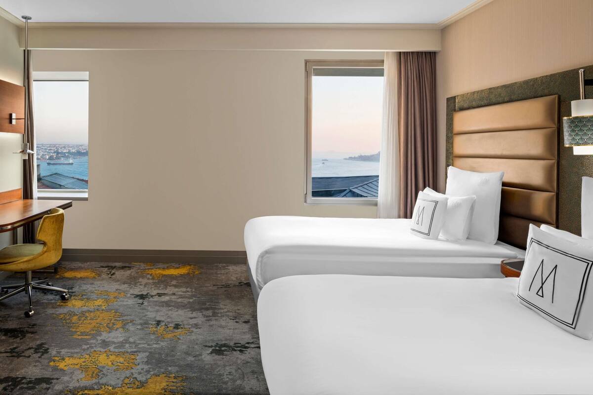 Een hotelkamer met twee bedden, een bureau, een stoel en een groot raam met uitzicht op de stad.
