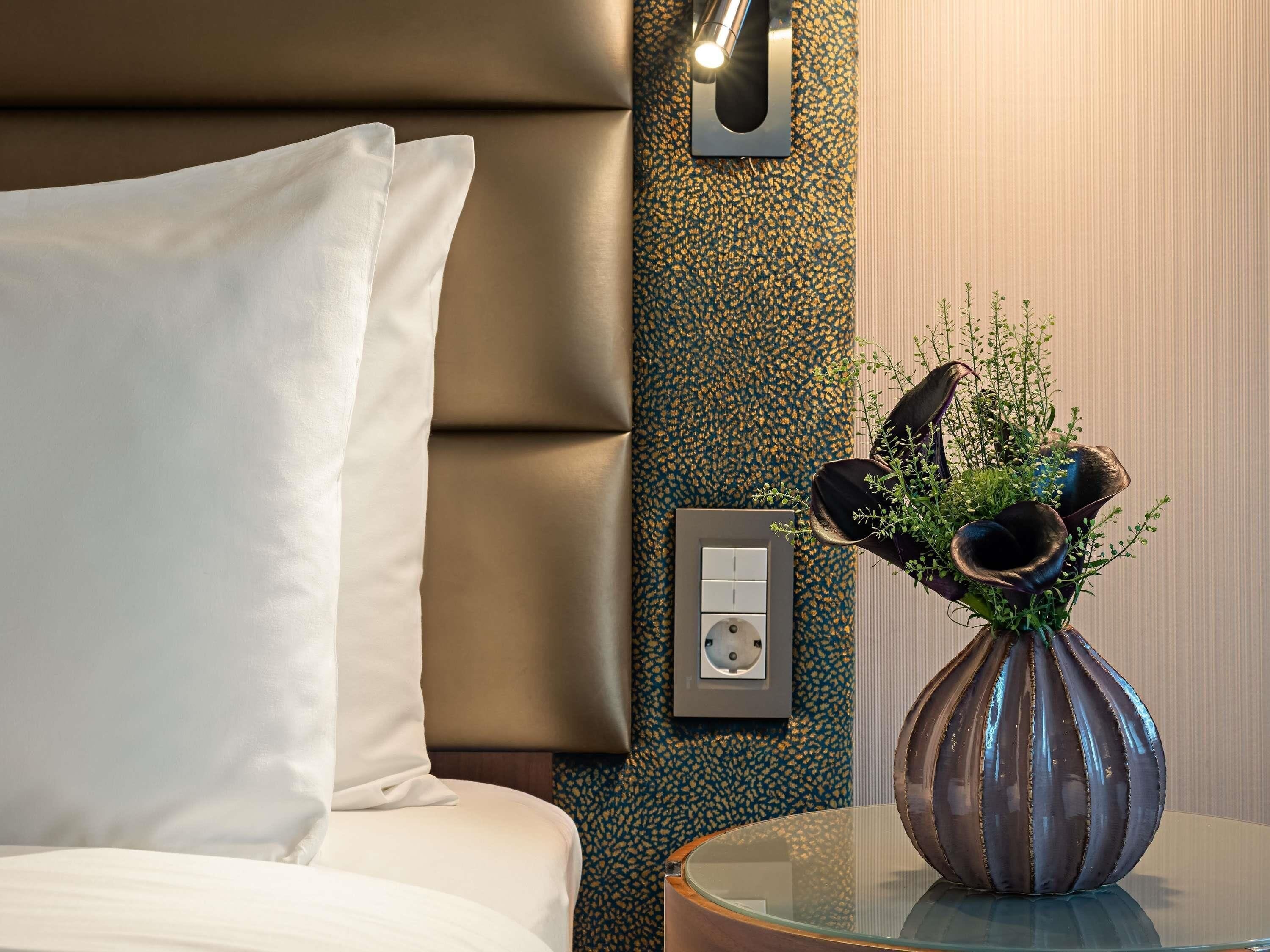 A hotel room with a bed, a wall-mounted light, a power outlet, and a vase with plants on a side table.