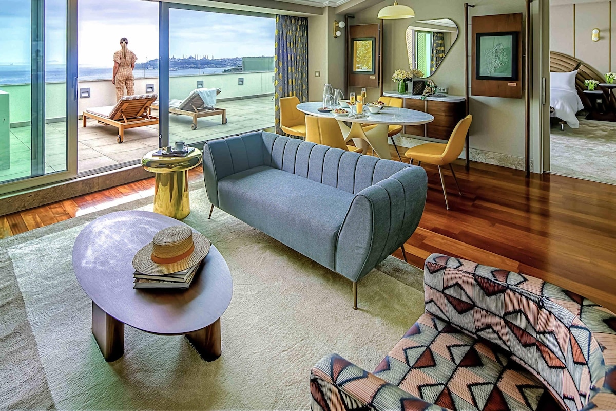 A modern hotel room with a large balcony, a dining area, and a living space with a sofa and a coffee table.