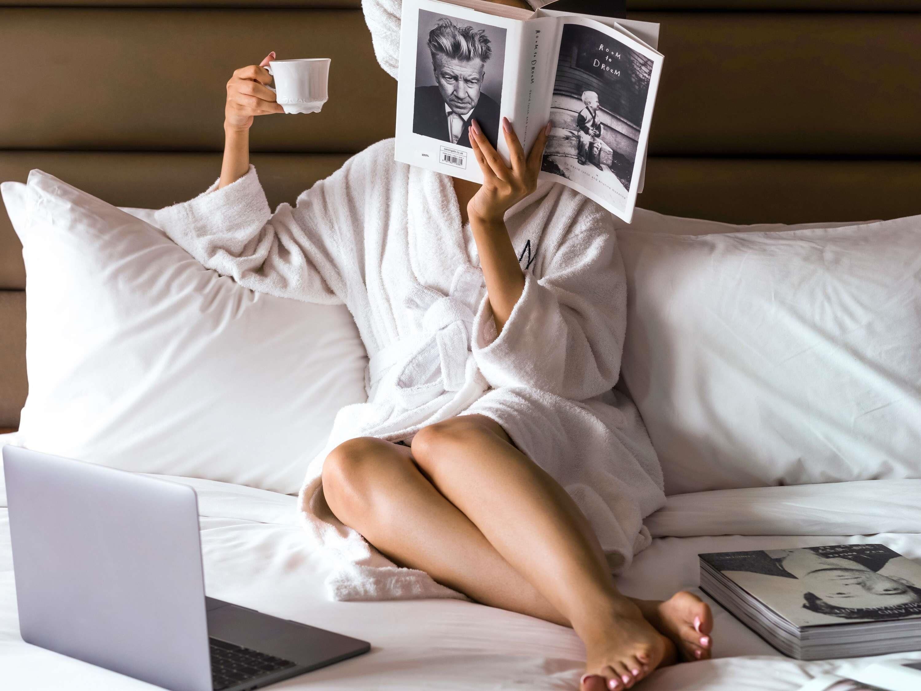 A person in a white bathrobe sitting on a bed with a laptop, holding a book and a cup.