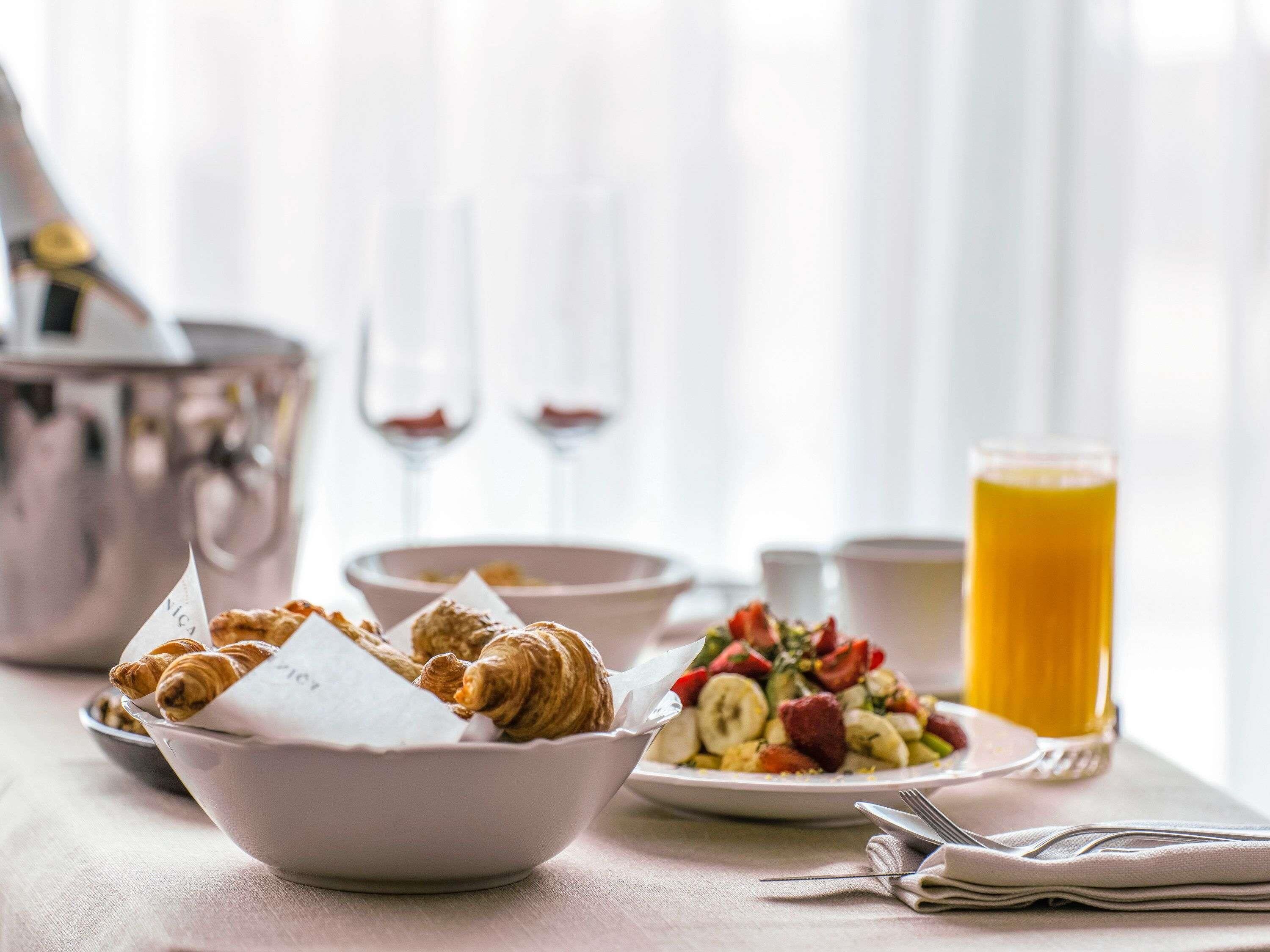 Une table dressée pour le petit-déjeuner, avec des croissants, une salade de fruits et un verre de jus d’orange.
