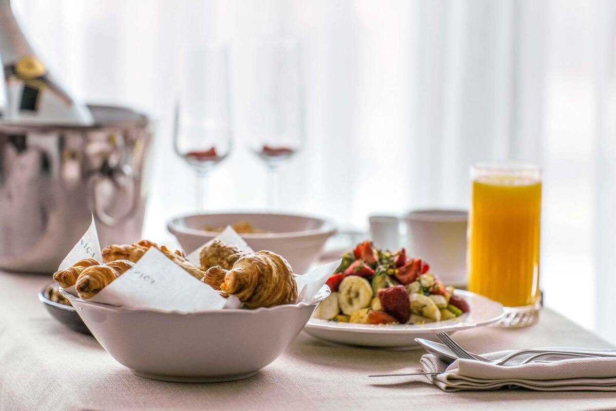 Une table dressée pour le petit-déjeuner, avec des croissants, une salade de fruits et un verre de jus d’orange.