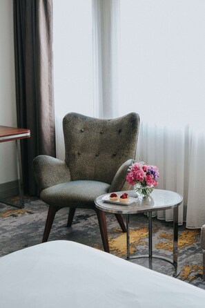 A hotel room with a grey upholstered armchair, a small round table with a vase of flowers, and a plate with pastries. - The Artisan Hotel Istanbul - MGallery Collection (Istanbul)