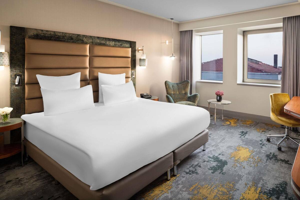 Een hotelkamer met een groot bed, een stoel, een klein tafeltje en uitzicht op de stad door het raam.