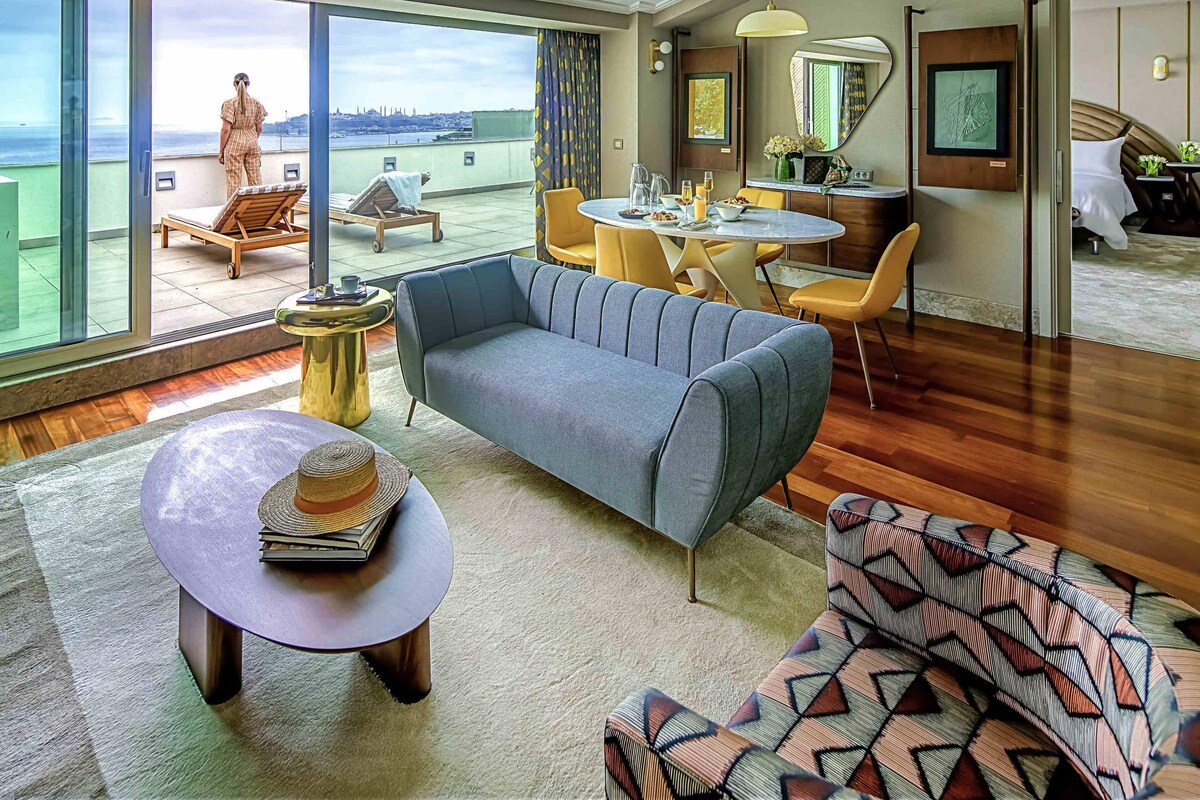 A modern hotel room with a large balcony, a dining area, and a living space with a sofa and a coffee table.