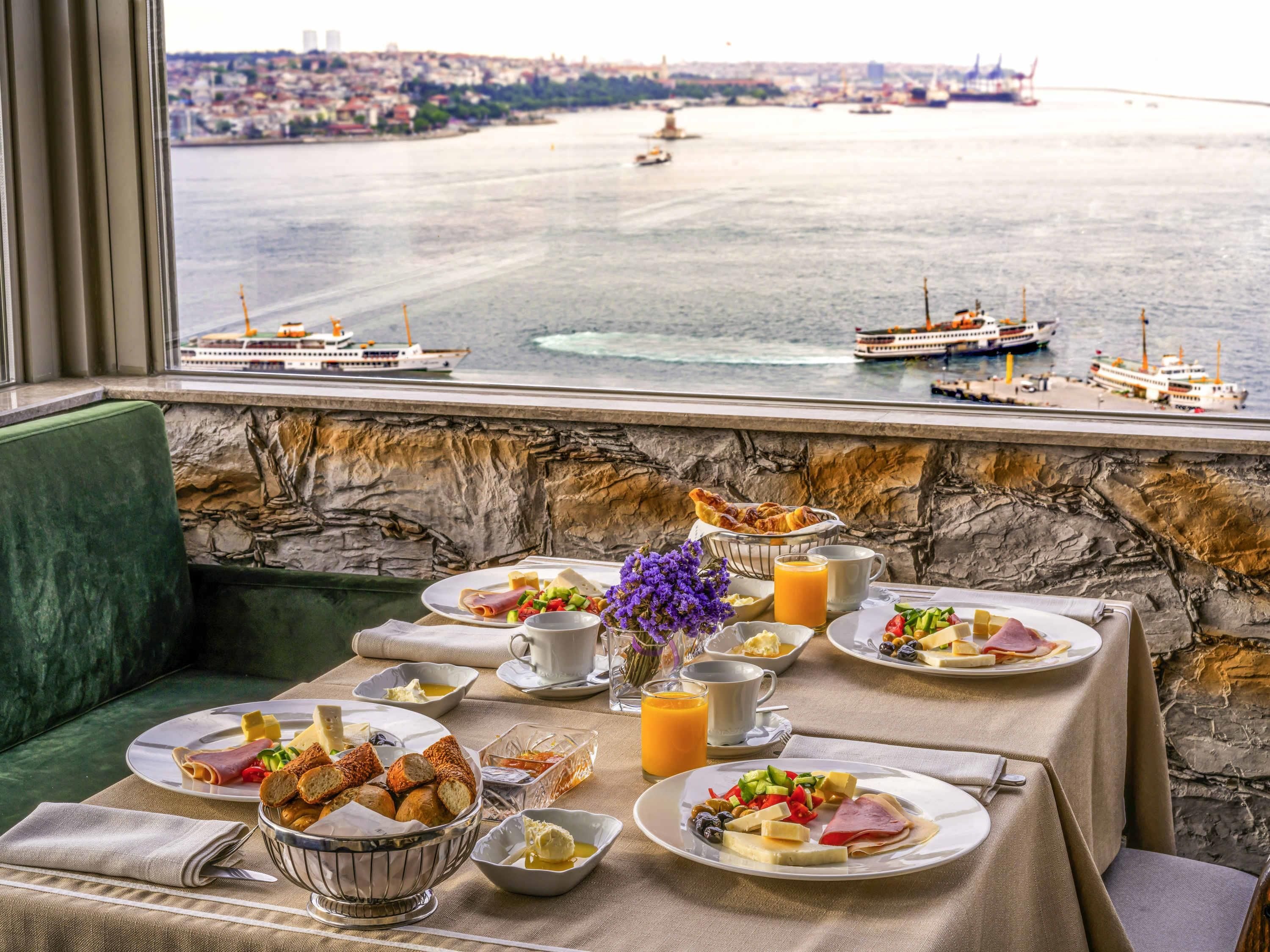 A breakfast setting with a view of the sea and boats, featuring plates of food, a vase of flowers, and a table with a stone wall backdrop.
