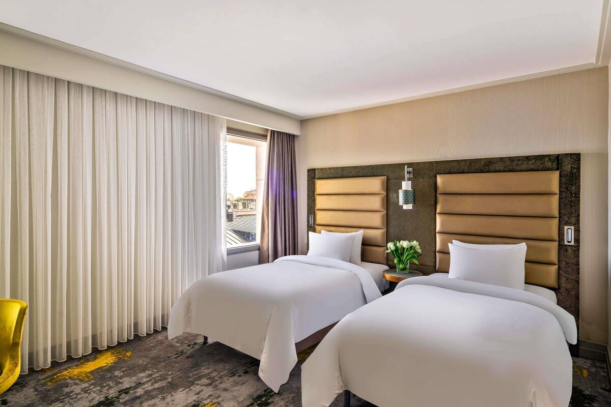 Een hotelkamer met twee bedden, een raam met gordijnen, een klein tafeltje met een vaas en een gele stoel.
