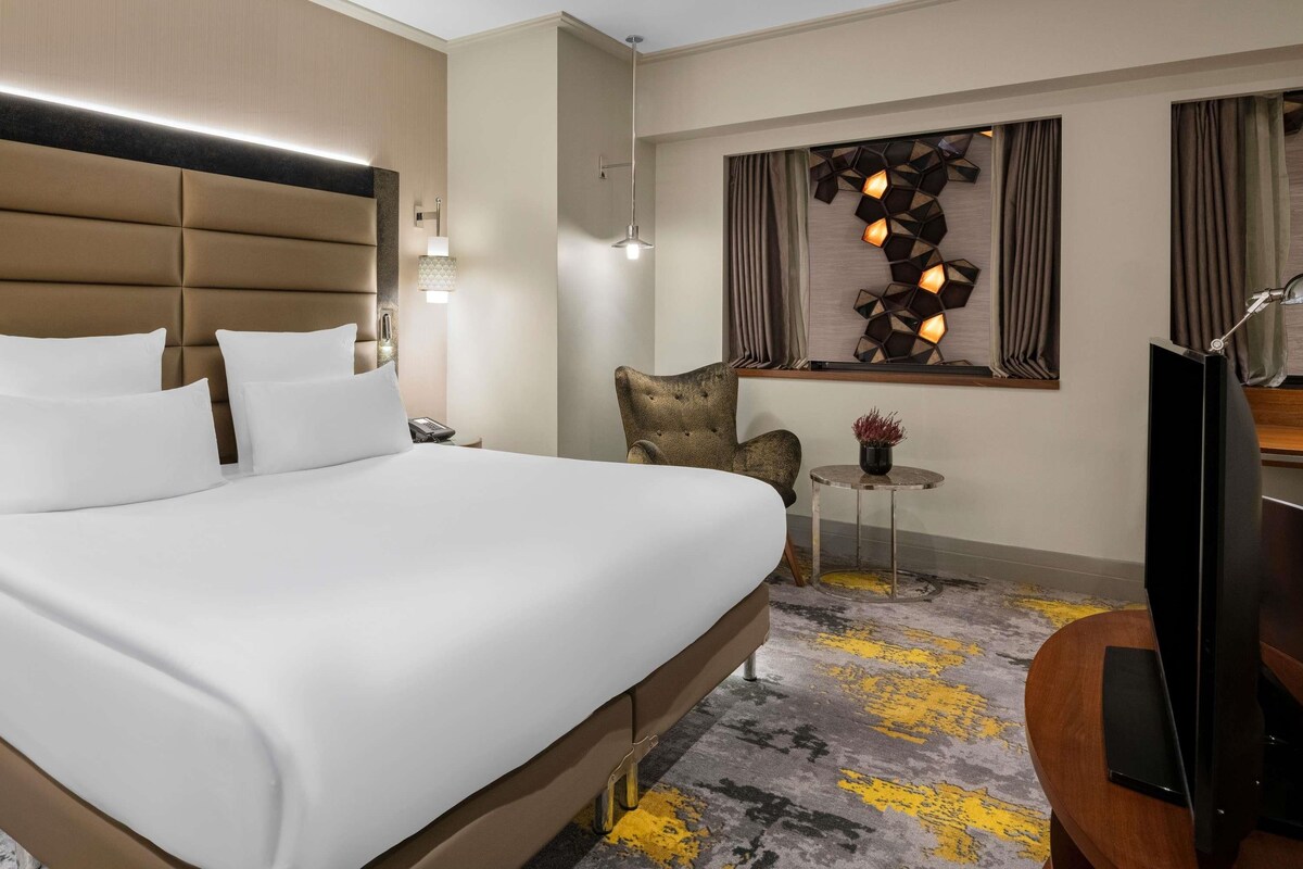 A hotel room with a large bed, a small table with a lamp, a chair, and a television.