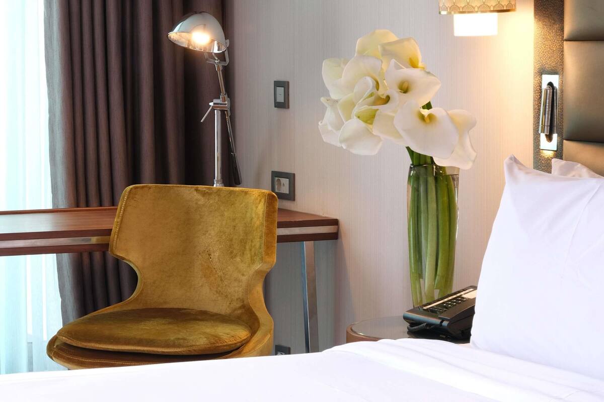 Een hotelkamer met een bed, een gouden fauteuil, een bureau met een lamp, een vaas met witte bloemen en een nachtkastje met een telefoon.