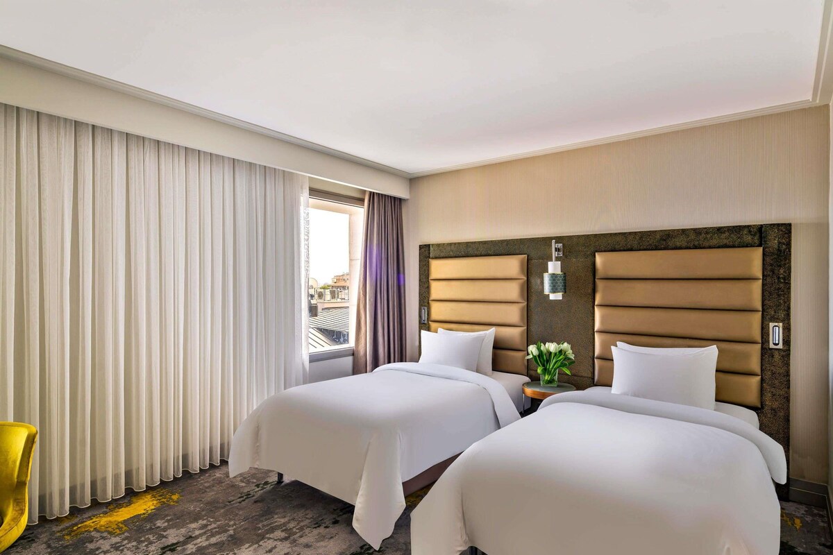 A hotel room with two beds, a window with curtains, a small table with a vase, and a yellow chair.