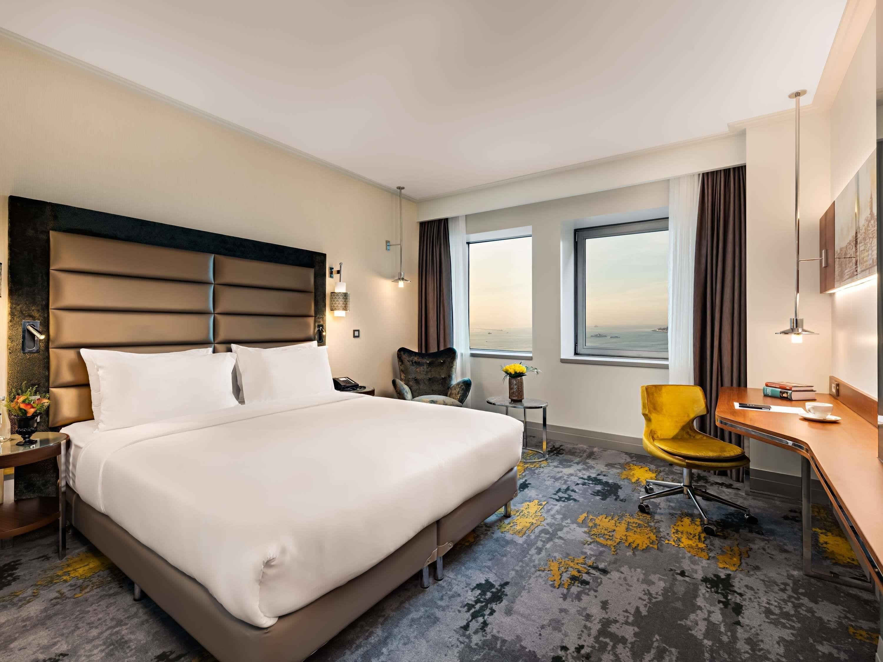 A hotel room with a large bed, a desk, a chair, and a view of the sea through the windows.