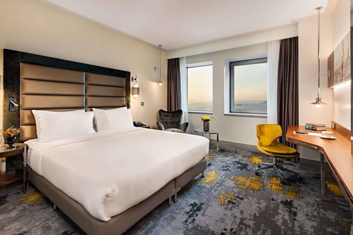 Een hotelkamer met een groot bed, een bureau, een stoel en uitzicht op de zee door de ramen.