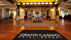Un hall d'entrée avec un comptoir de réception, une table centrale entourée de chaises et un tapis à motifs portant l'inscription « Grand Casino Hotel Resort ».
