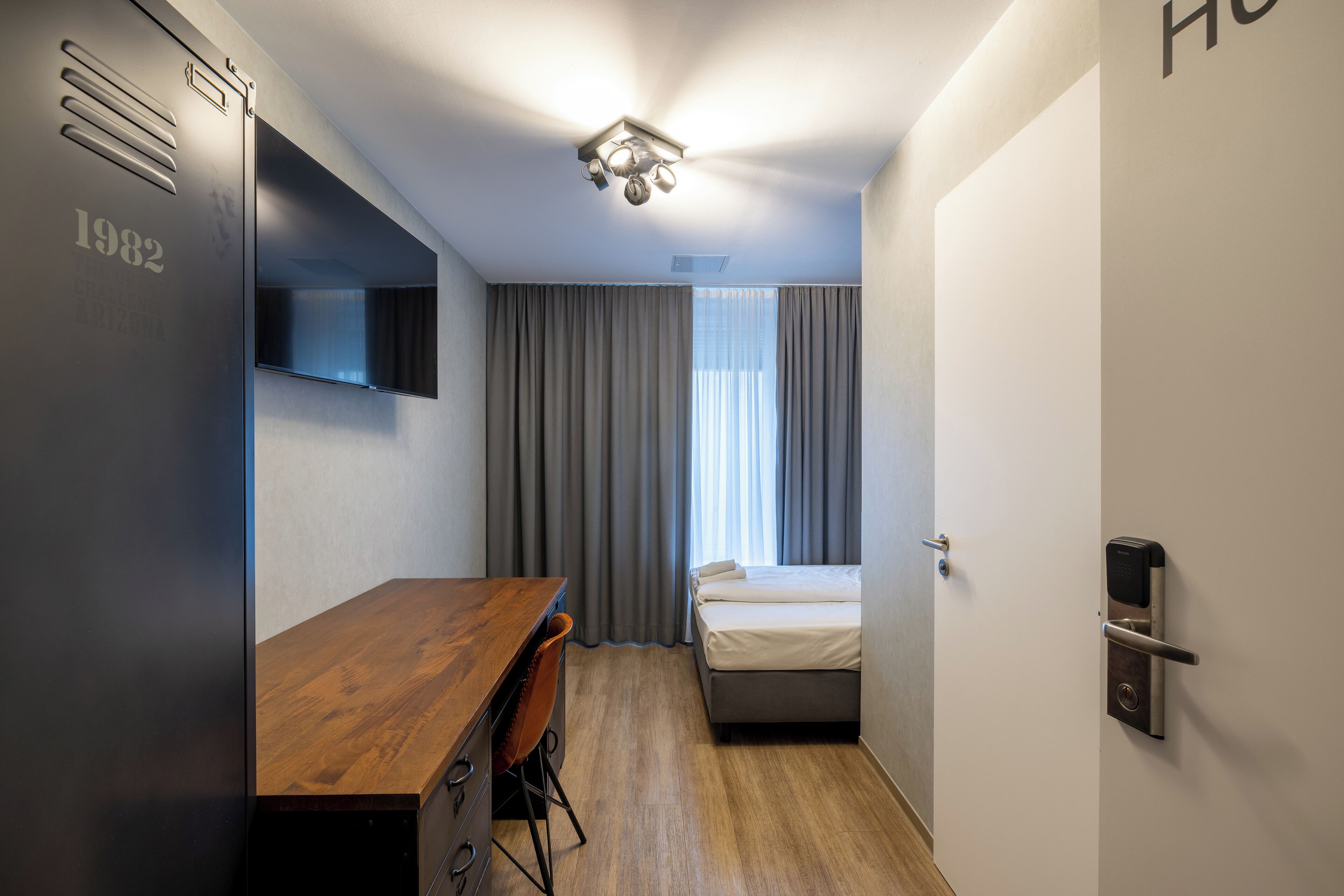 Une chambre d’hôtel compacte, dotée d’un bureau en bois, d’un lit, d’une armoire et d’un luminaire moderne au plafond.