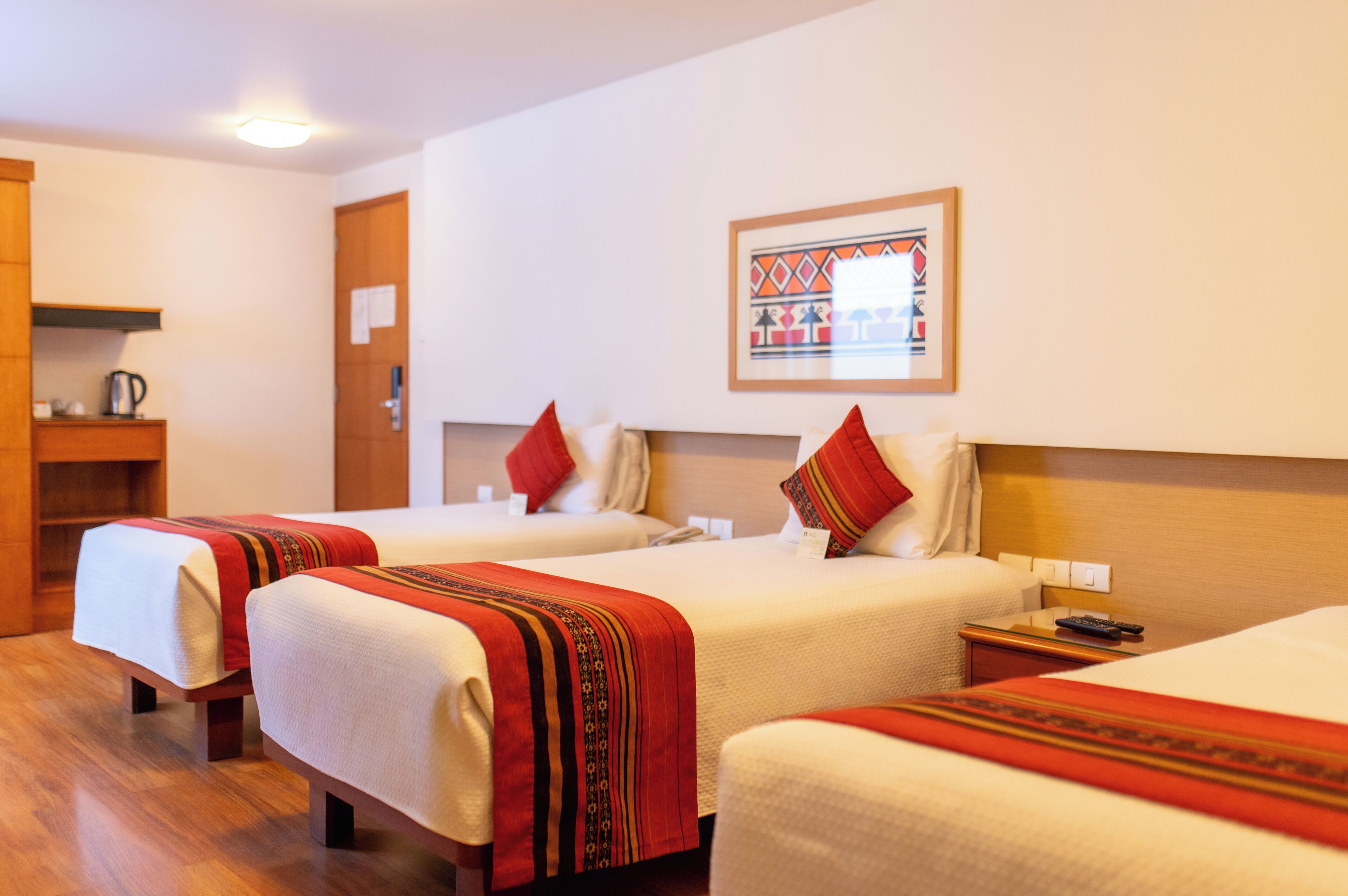 A hotel room with two beds, each with a red and white blanket, a wooden headboard, a bedside table with a remote control, and a wall-mounted picture.