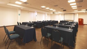 Un salón de conferencias con varias mesas y sillas, equipado con botellas de agua y vasos, y una pantalla de proyector.