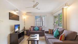 Uma sala de estar com dois sofás, uma mesa de centro, uma televisão em um móvel, um ventilador de teto e uma janela com persianas.