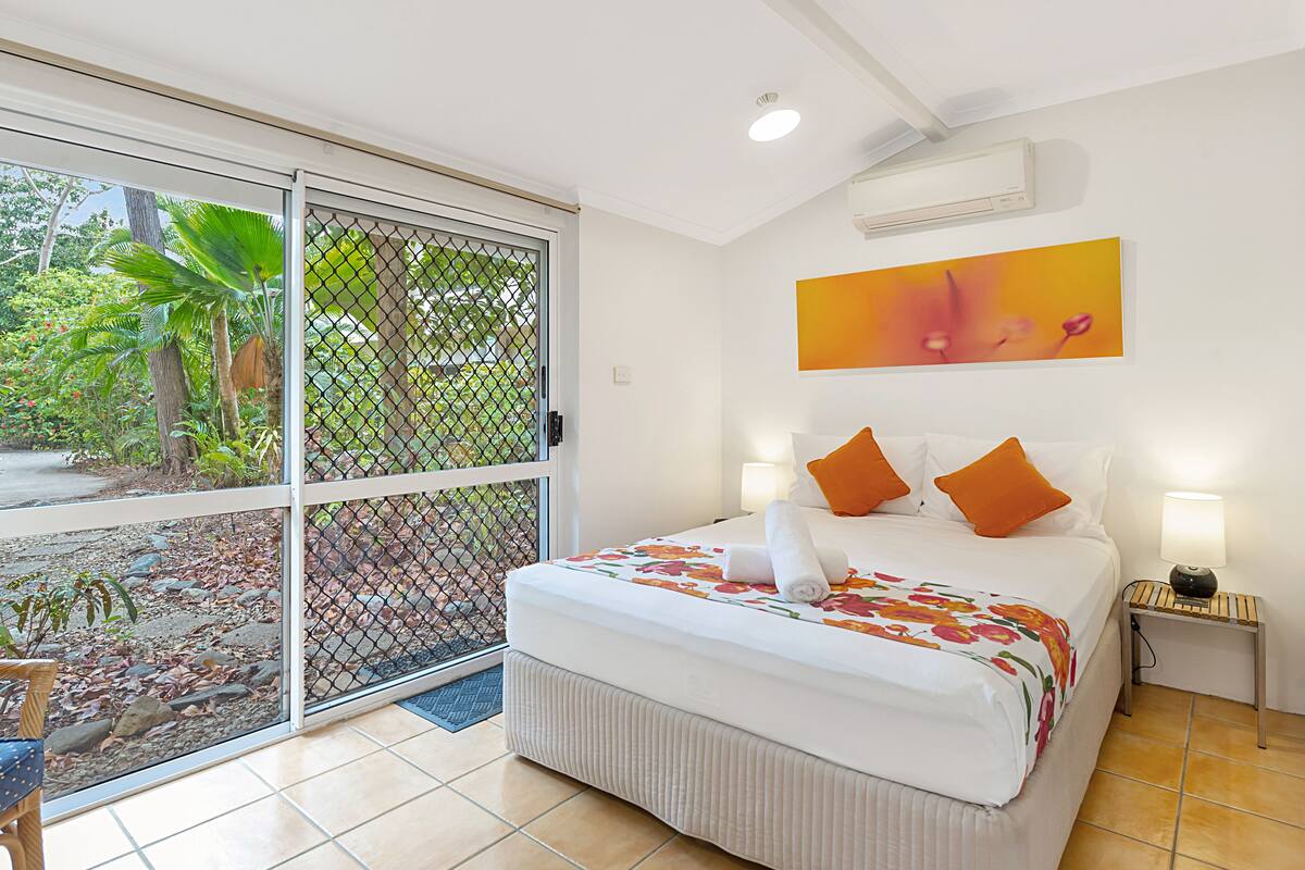 A hotel room with a bed, a nightstand, a window with a view of greenery, and a ceiling fan.