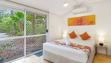 A hotel room with a bed, a nightstand, a window with a view of greenery, and a ceiling fan.