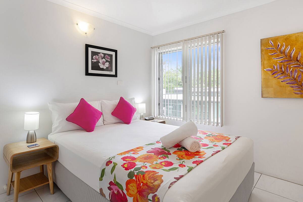A hotel room with a bed, a nightstand, a lamp, a framed picture, a window with blinds, and a painting on the wall.