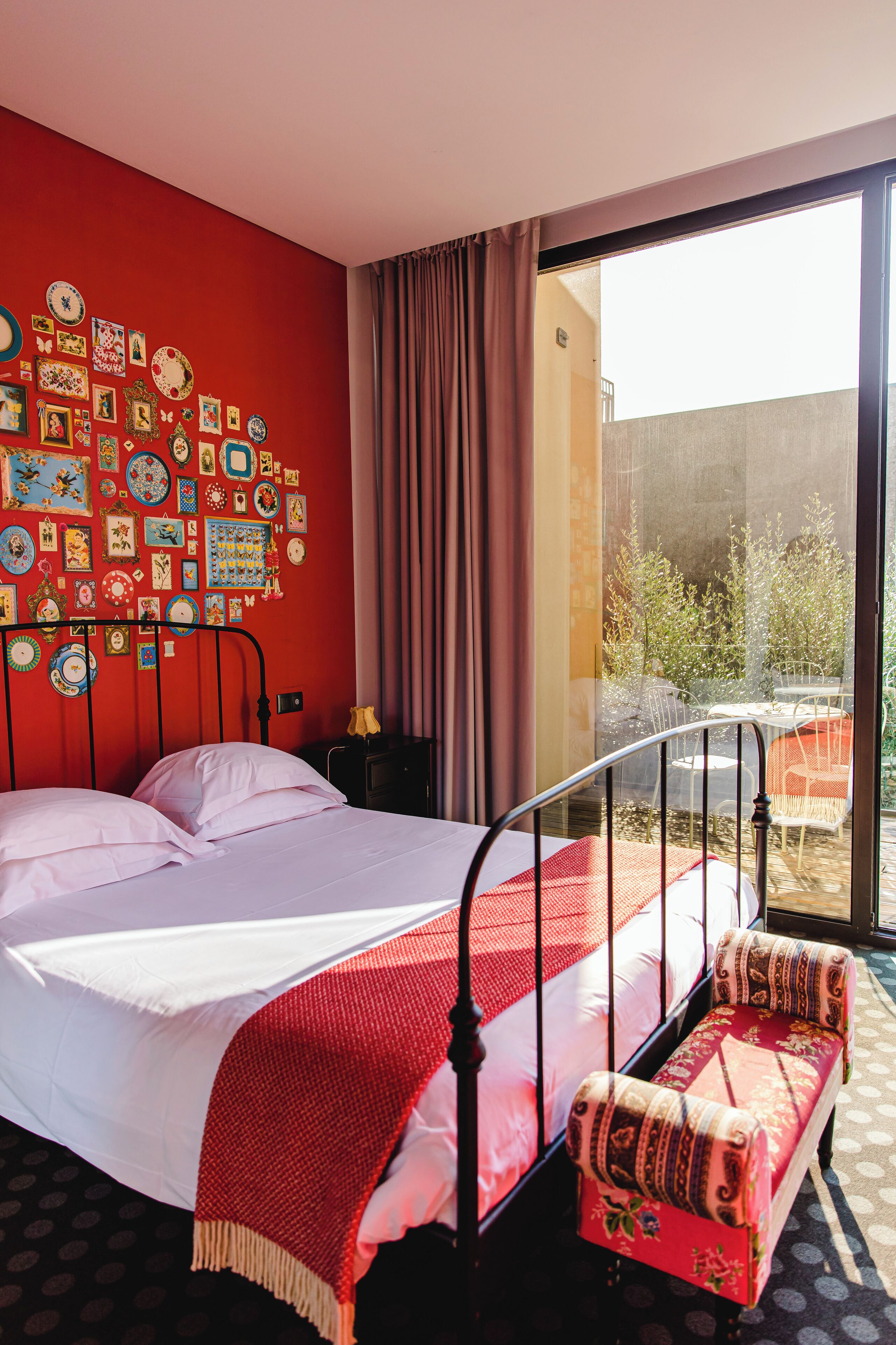 Une pièce avec un lit, un banc, un mur rouge orné de tableaux et une vue sur l’extérieur.