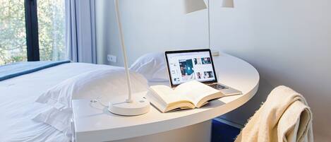 A modern hotel room with a bed, a desk with a laptop and book, a chair with a blanket, and a large window with curtains.