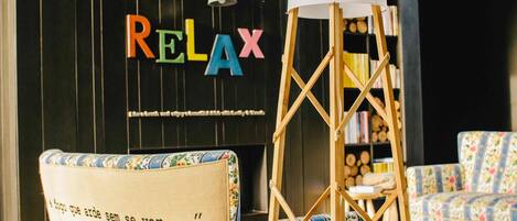 A room with a black wall featuring colorful 'RELAX' letters and a quote by Luis de Camões. There is a wooden easel with a white lampshade and a floral-patterned chair.