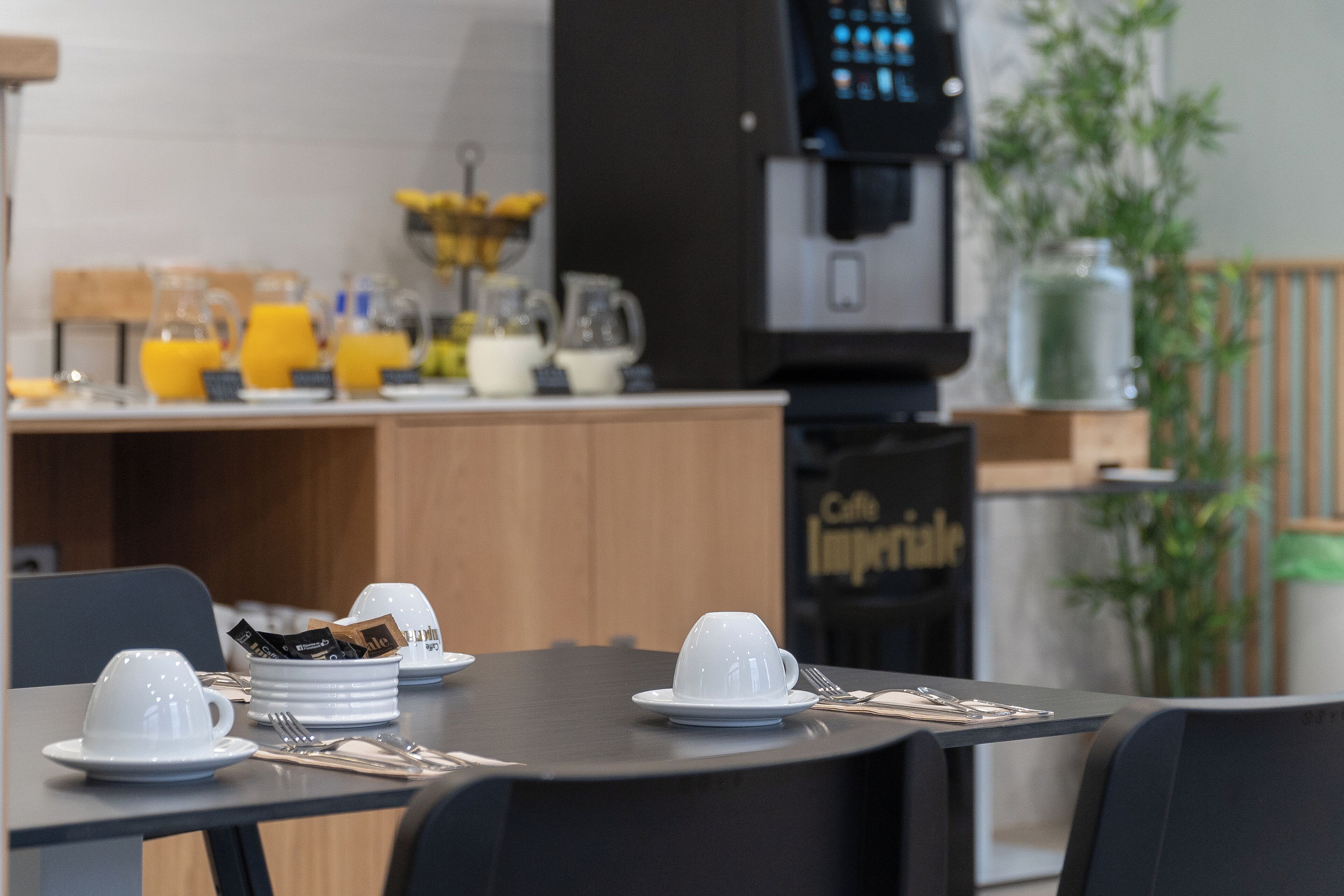 Ein Frühstückstisch mit weißen Tassen und Untertassen, einer Kaffeemaschine und einem Obstkorb.