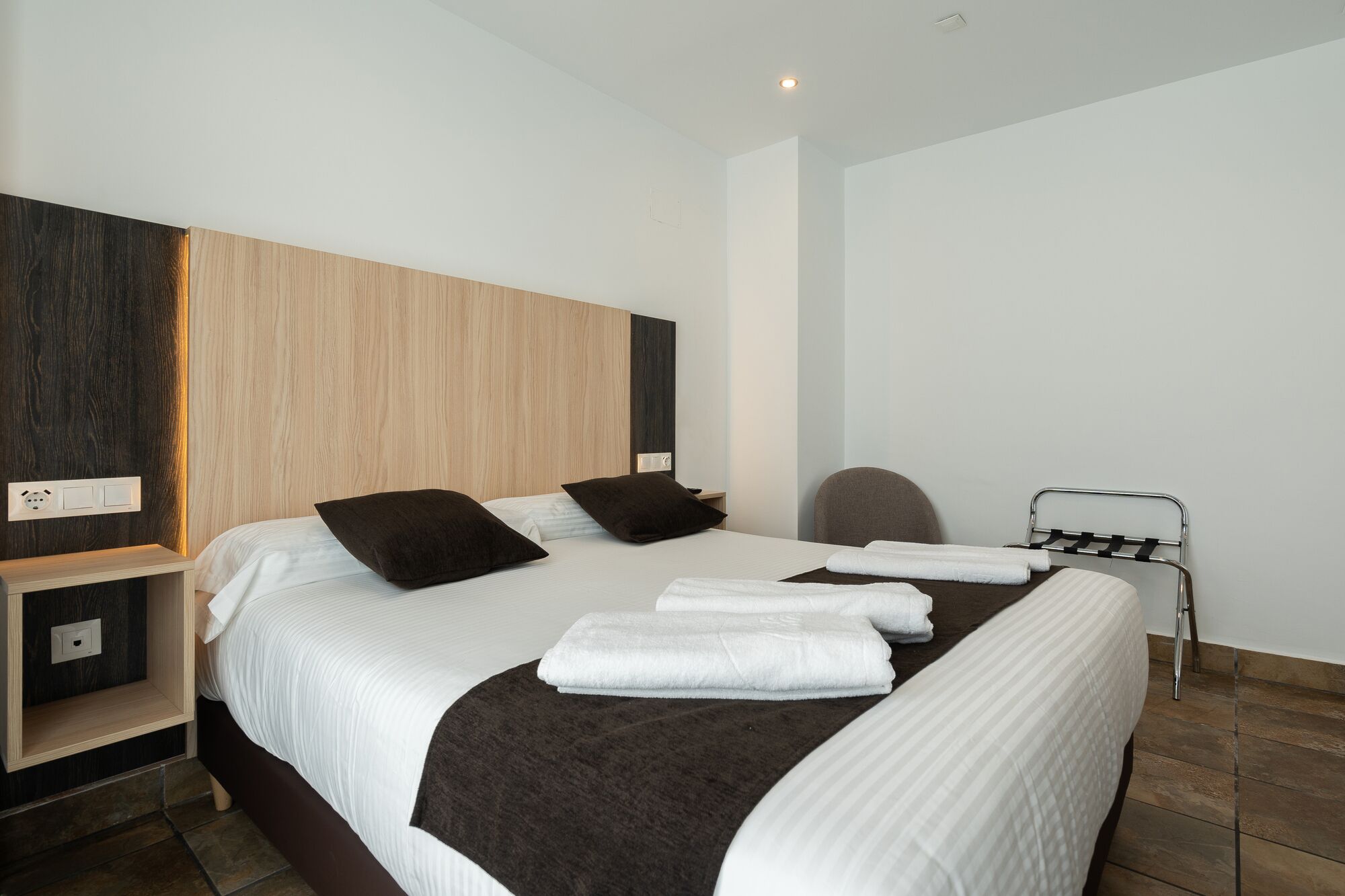 Ett modernt hotellrum med en stor säng, ett sängbord, en stol och en handduksställning.
