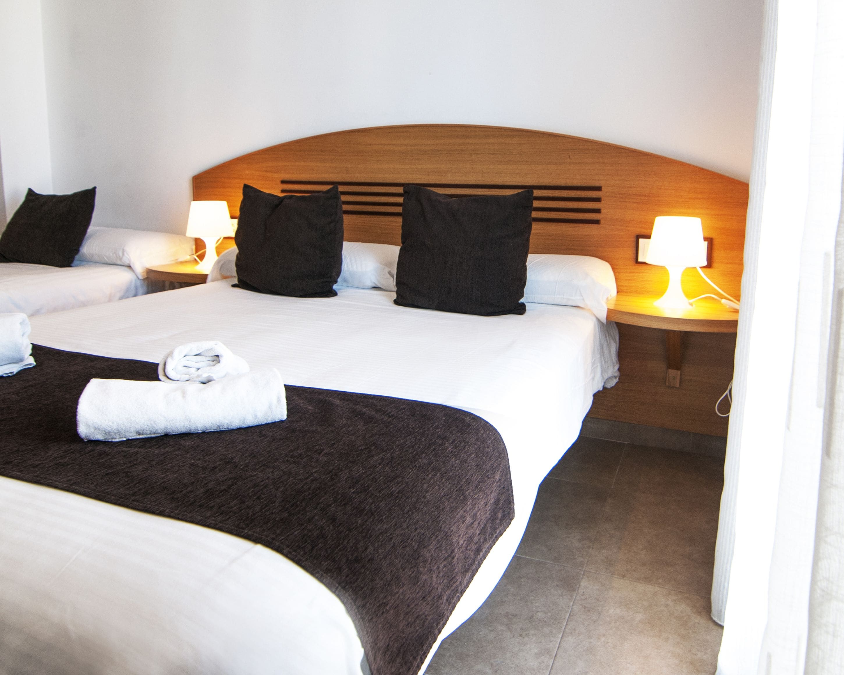 Ein Hotelzimmer mit zwei Betten, jeweils mit einem hölzernen Kopfteil, weißen Laken und schwarzen Kissen. An den Betten stehen Nachttischlampen und ein gefaltetes Handtuch.