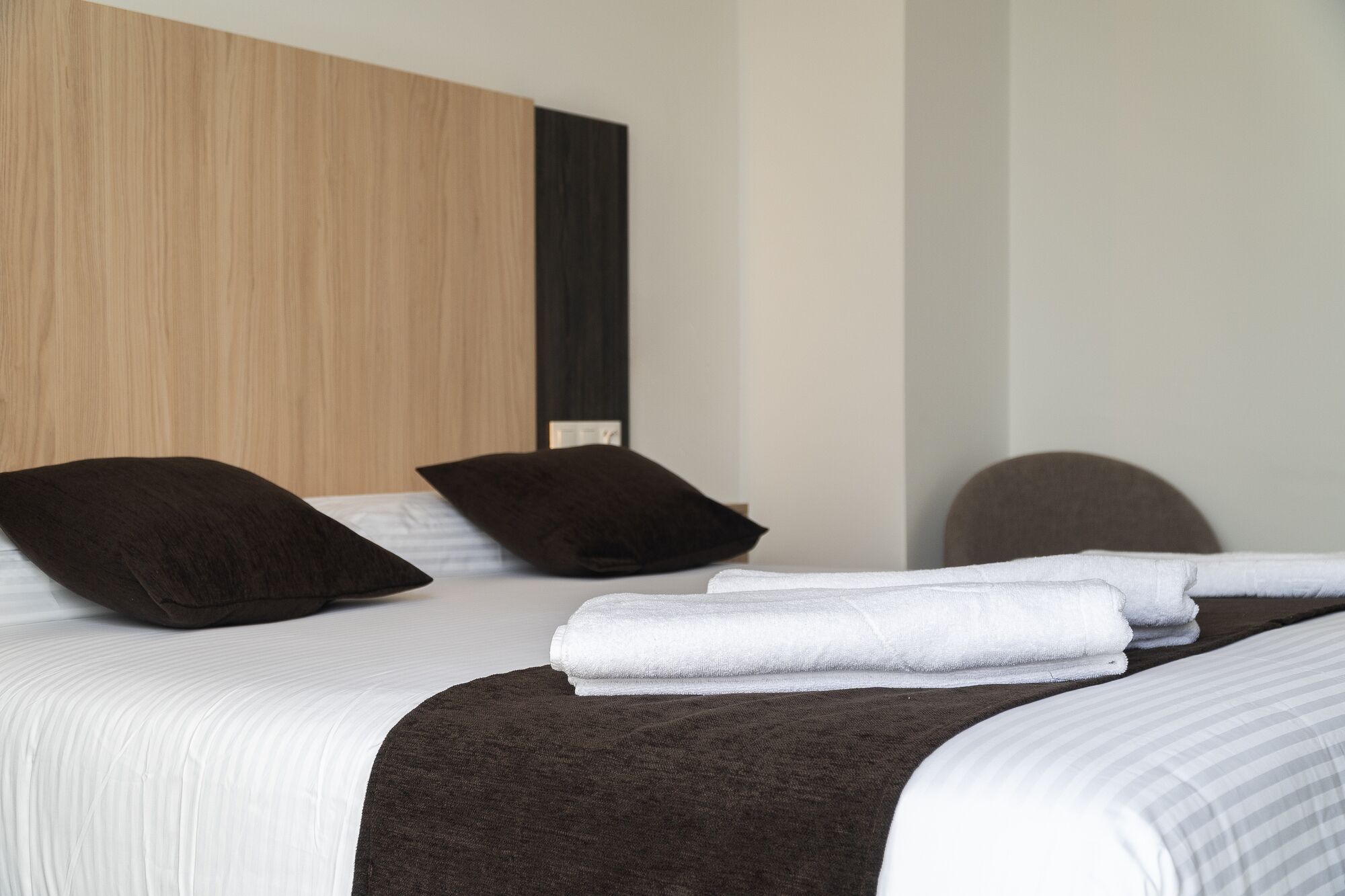 A hotel room with a bed, two dark pillows, a white blanket, and a folded white towel.