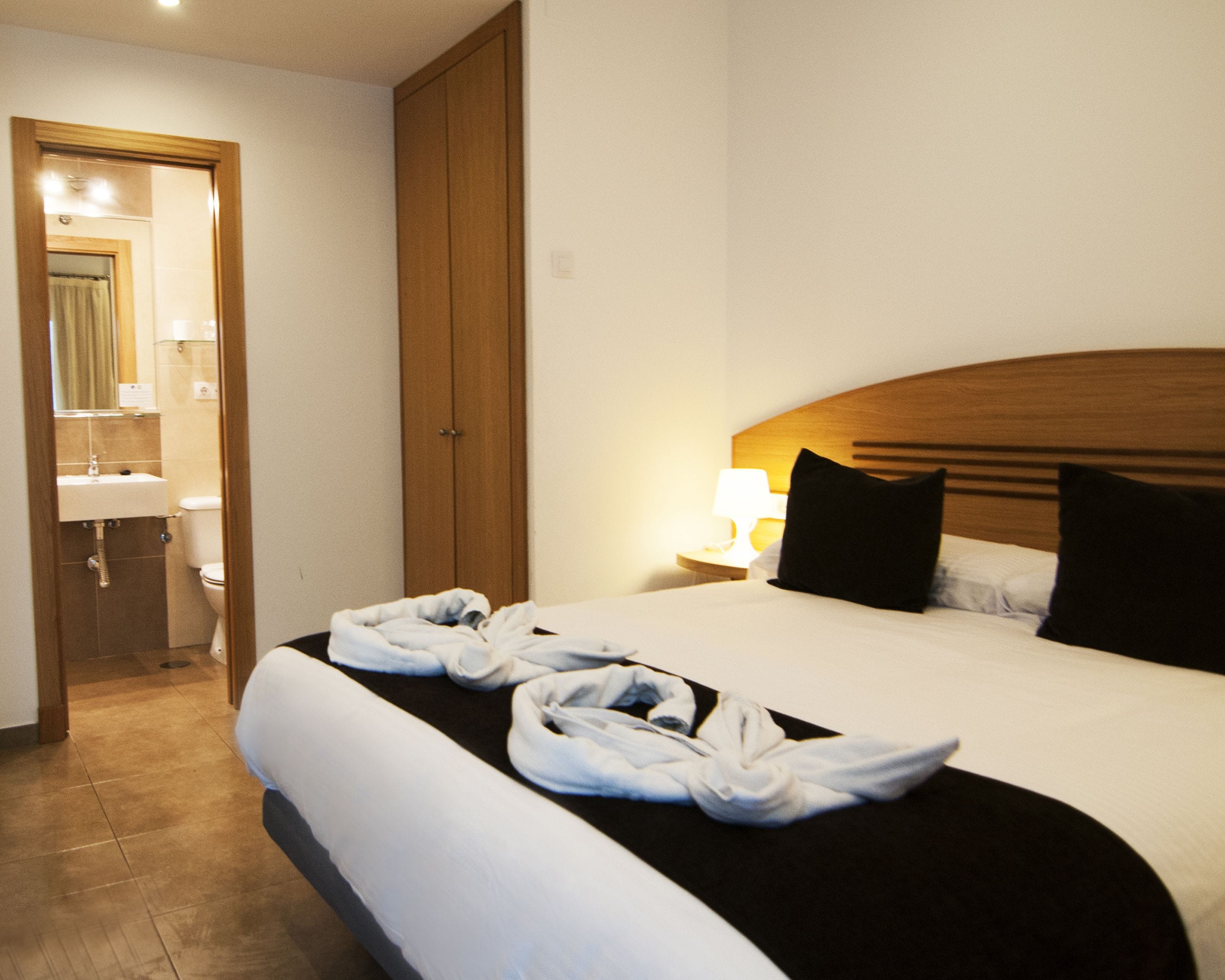 A hotel room with a bed, a nightstand with a lamp, a wooden wardrobe, and a bathroom visible through an open door.