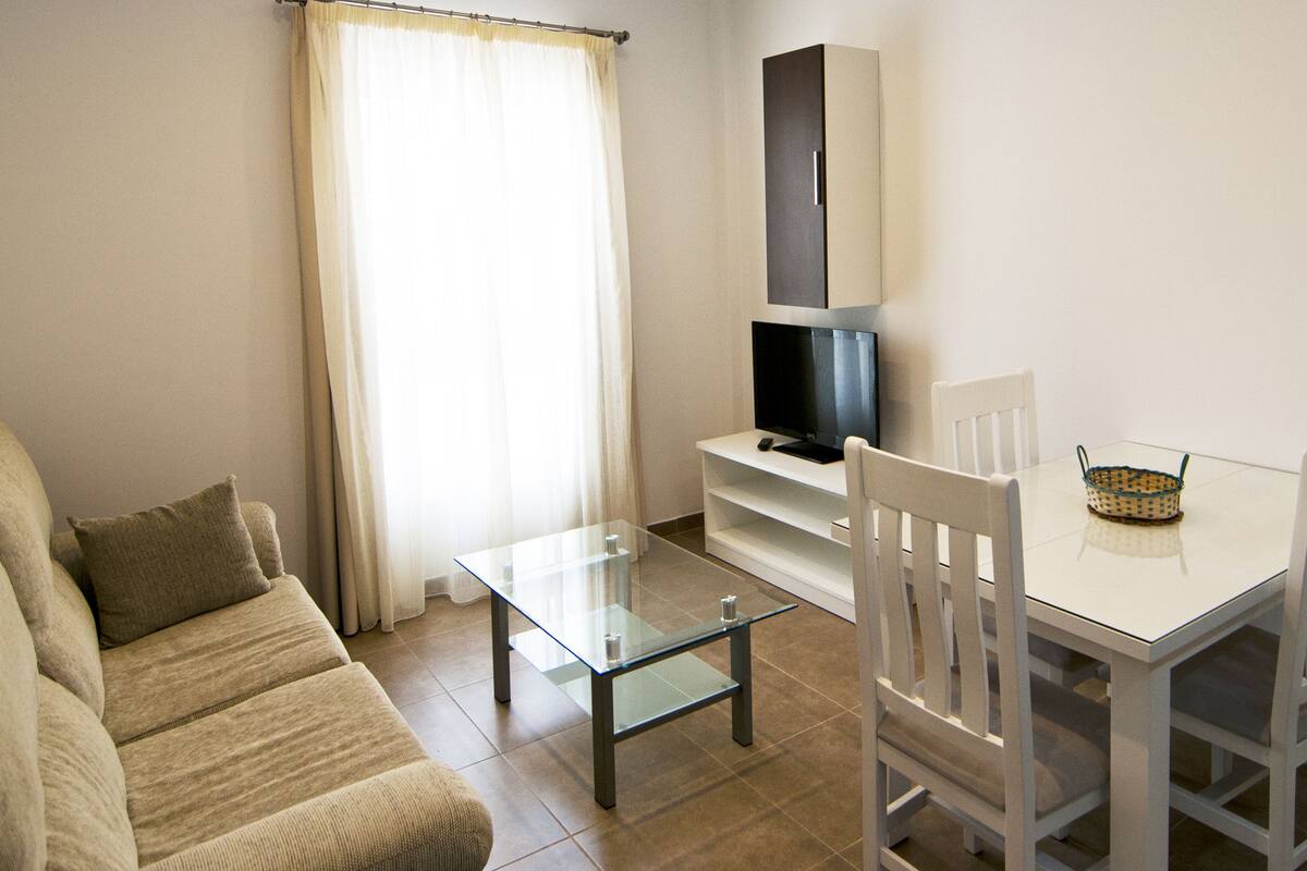 Un salon comprenant un canapé, une table basse en verre, un téléviseur posé sur un meuble et une table à manger avec des chaises.