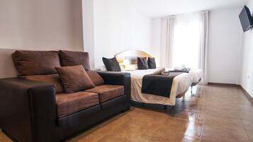 A hotel room with a brown sofa, a bed with a white headboard, and a flat-screen TV mounted on the wall.