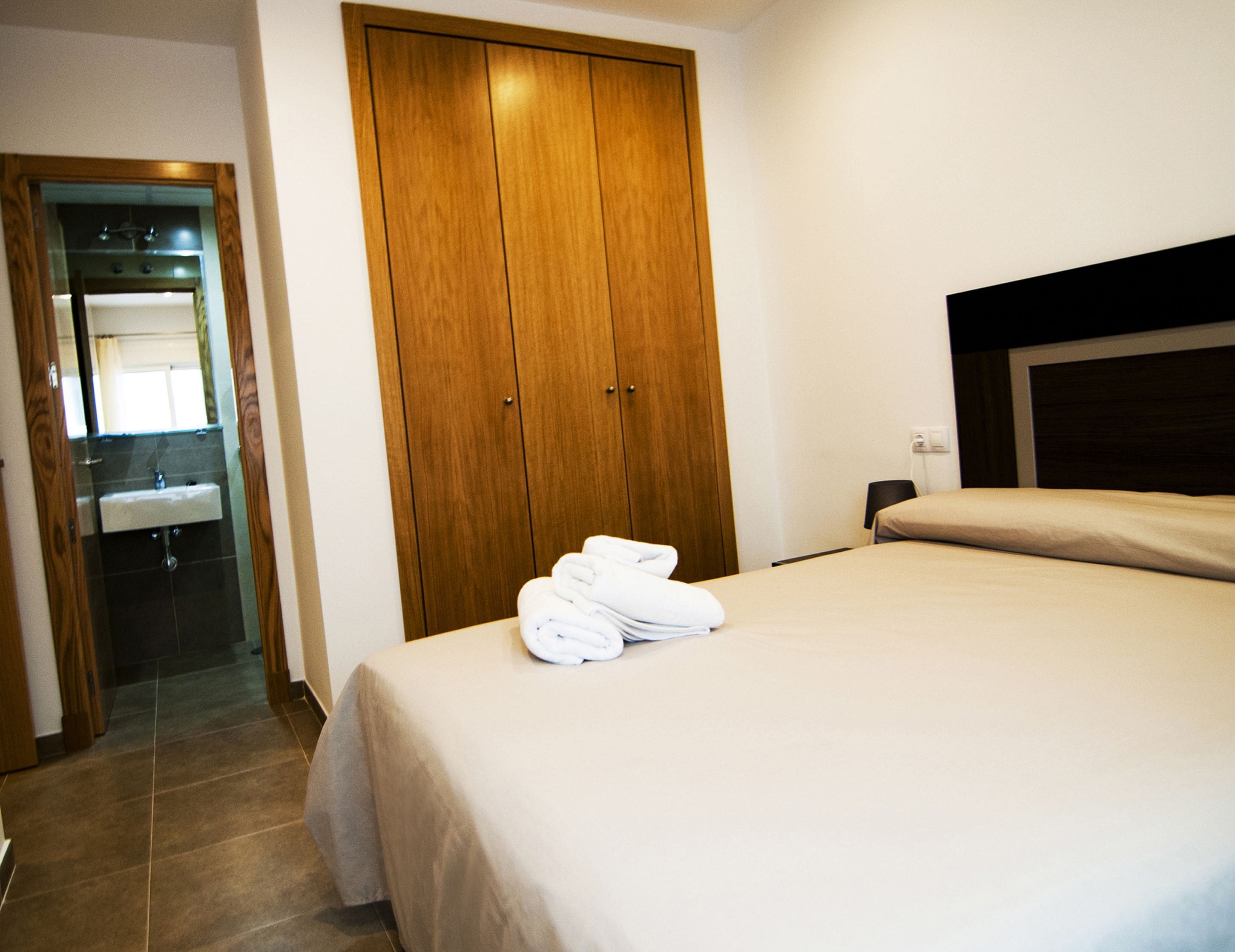A hotel room with a bed, a wooden wardrobe, a bathroom with a sink, and a mirror.
