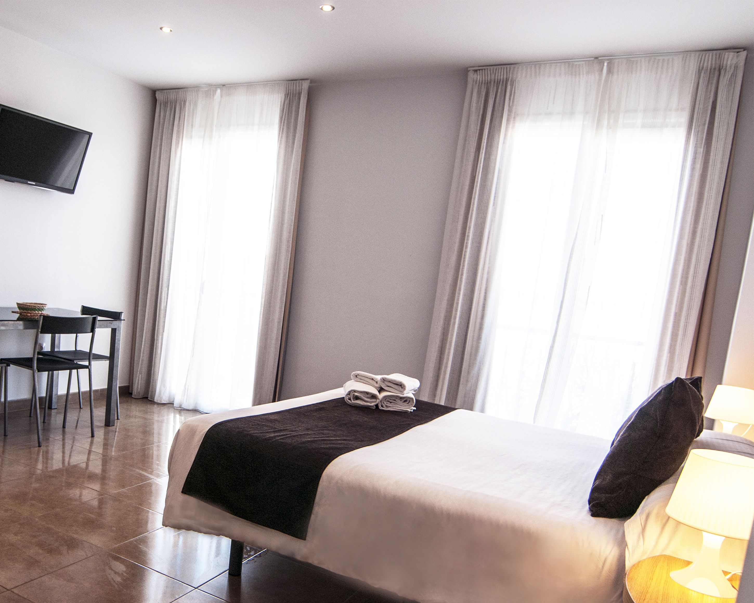 A hotel room with a bed, a TV mounted on the wall, a desk with chairs, and large windows with curtains.