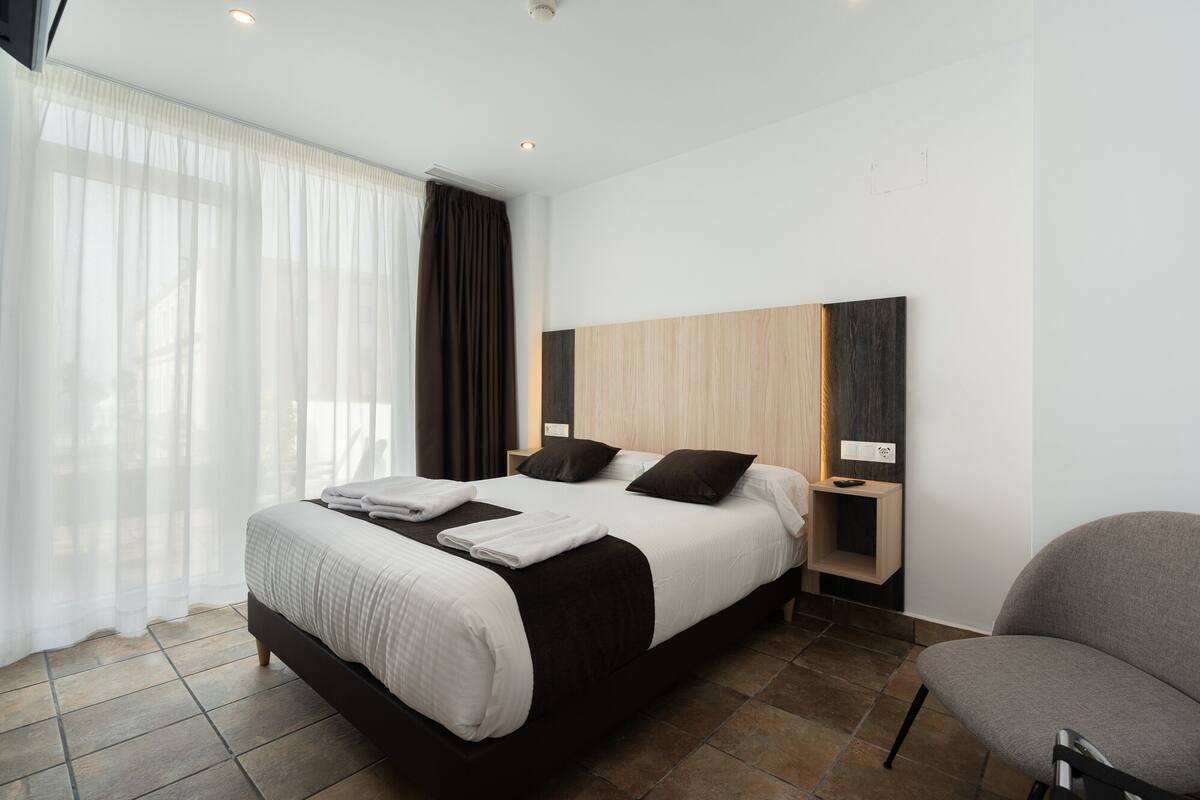 Une chambre d’hôtel moderne, dotée d’un grand lit, d’une chaise, d’une table de chevet et d’une fenêtre avec des rideaux légers.